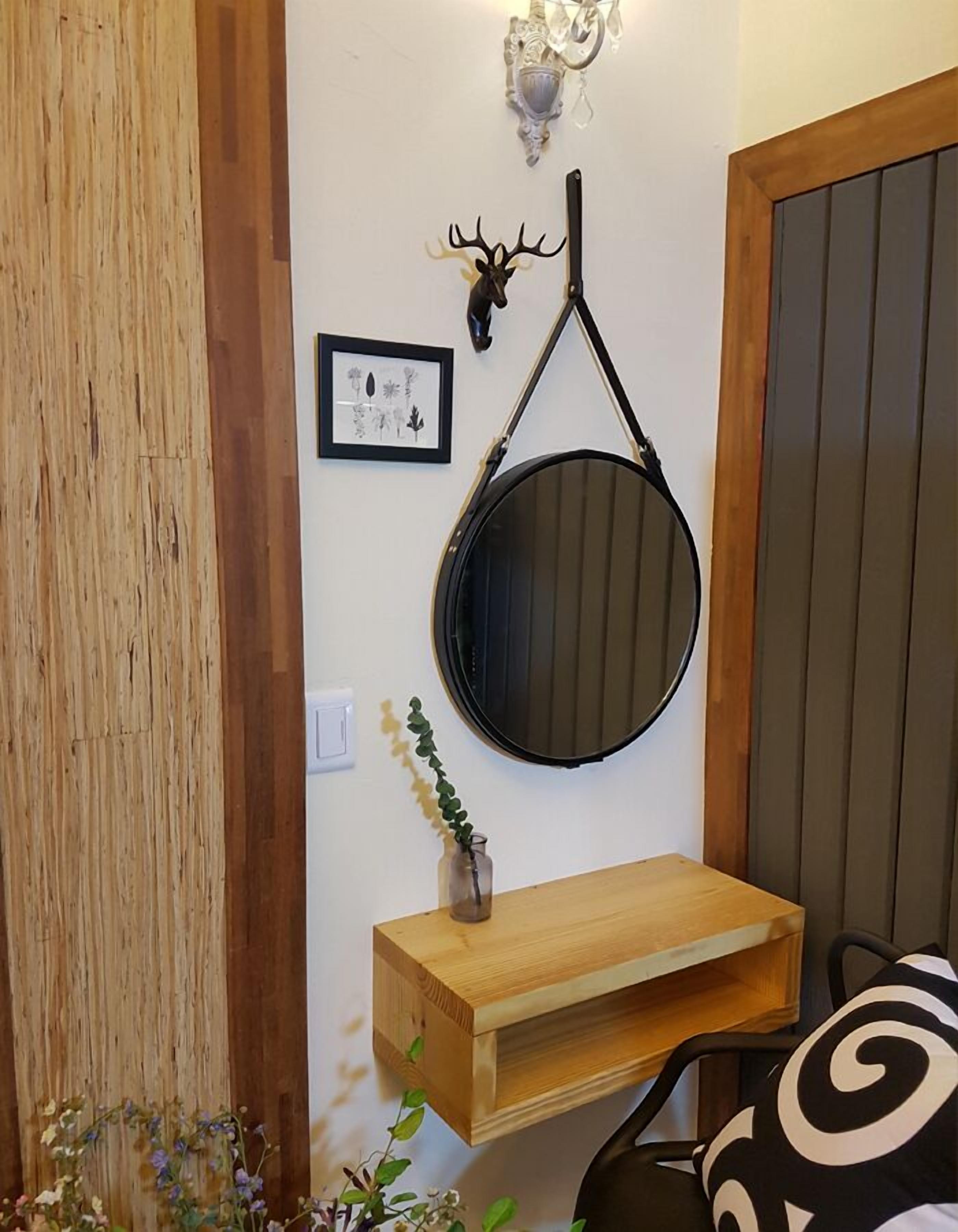 Ett rum med en väggpanel i trä, ett upphängt rådjurshuvud, en inramad botanisk illustration, en rund spegel och en liten trähylla med en växt.