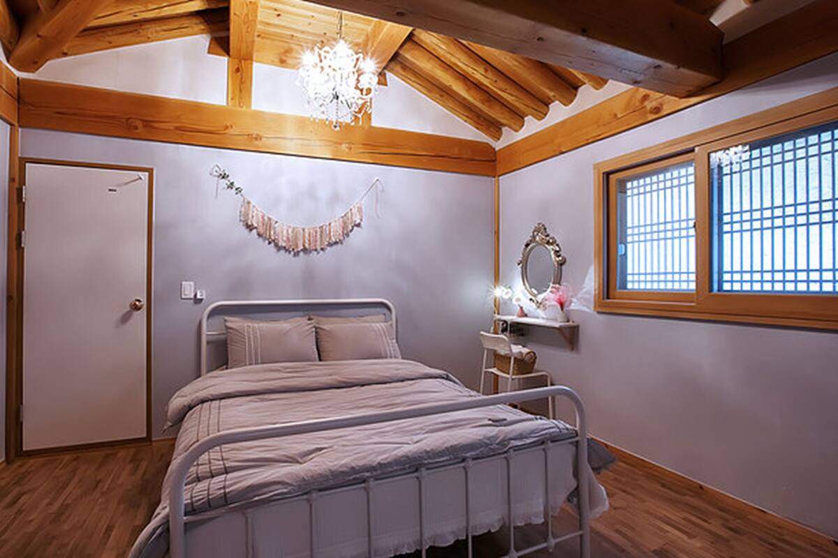 Ein Schlafzimmer mit Bett, Holzbalkendecke, einer Chandelieber, einem Fenster mit Gitterstäben und einer Tür.