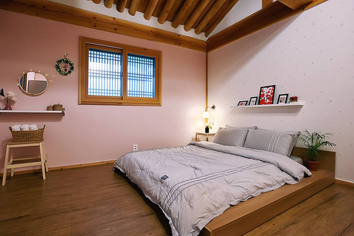 Ein Schlafzimmer mit einem hölzernen Bett, einem kleinen Nachttisch, einem Regal mit Dekoration und einer Zimmerpflanze. Es gibt außerdem einen Spiegel und ein Fenster mit Gitterstäben.