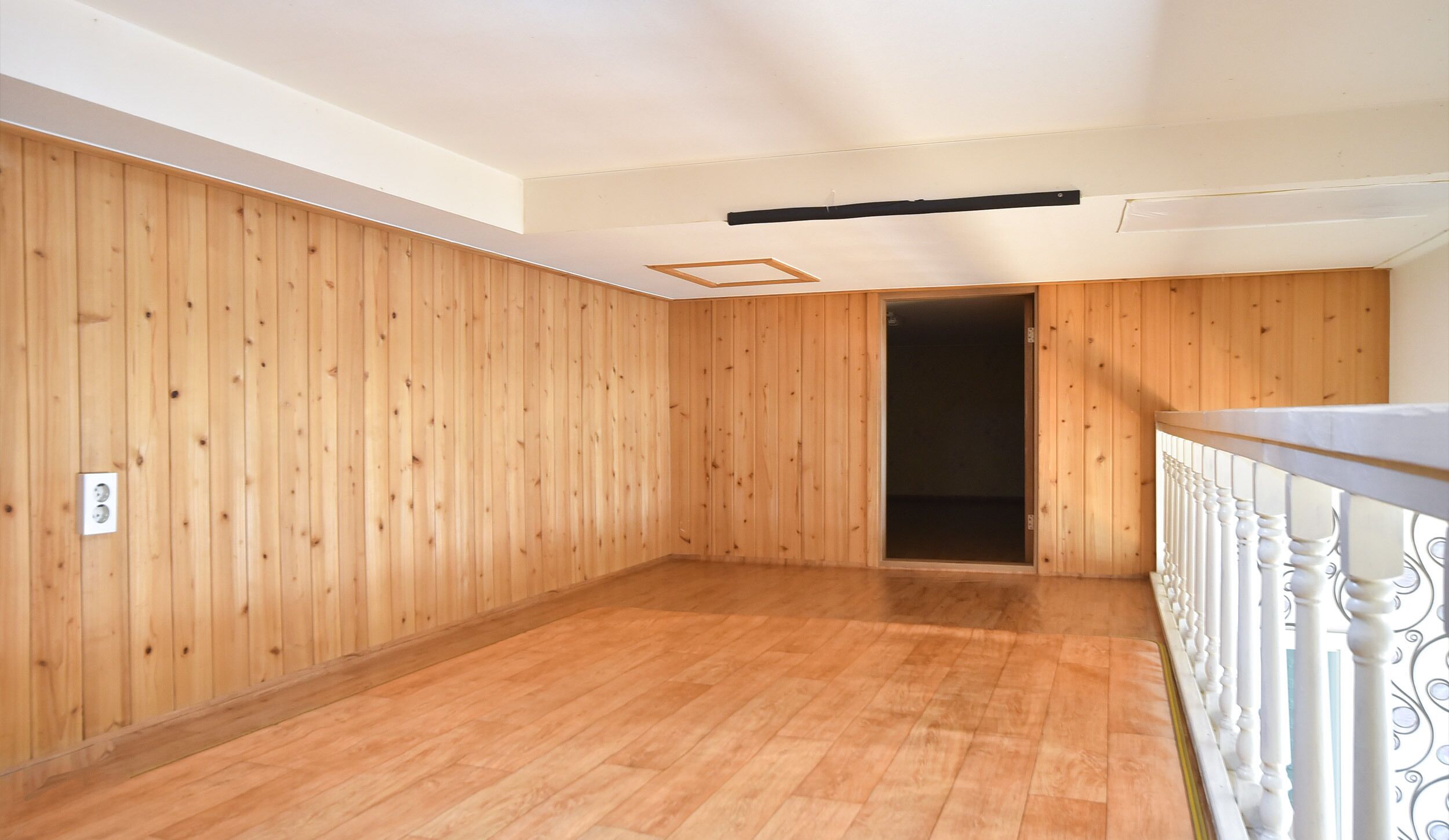 A room with wooden panel walls, a wooden floor, a white railing, and a door leading to another room.