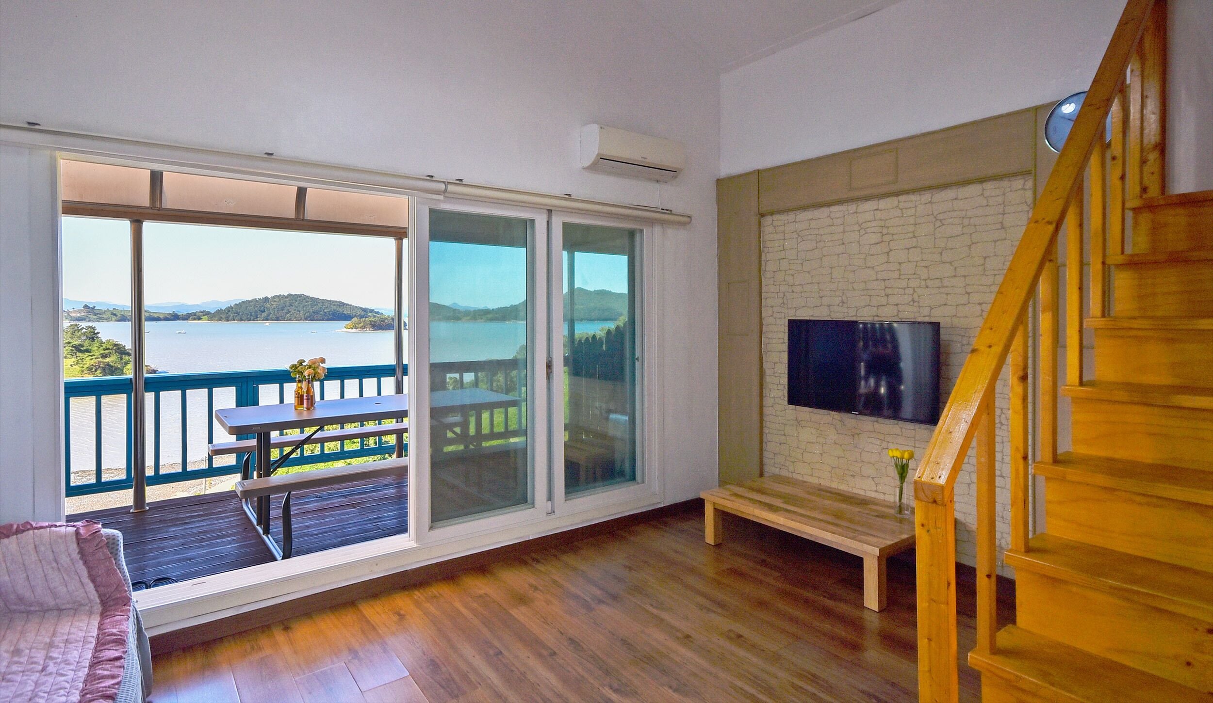 A room with a large sliding glass door leading to a balcony with a view of a lake and mountains. There is a wooden bench and a small table with a vase of flowers on the balcony.