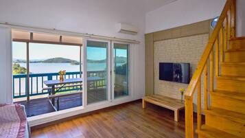 A room with a large sliding glass door leading to a balcony with a view of a lake and mountains. There is a wooden bench and a small table with a vase of flowers on the balcony.