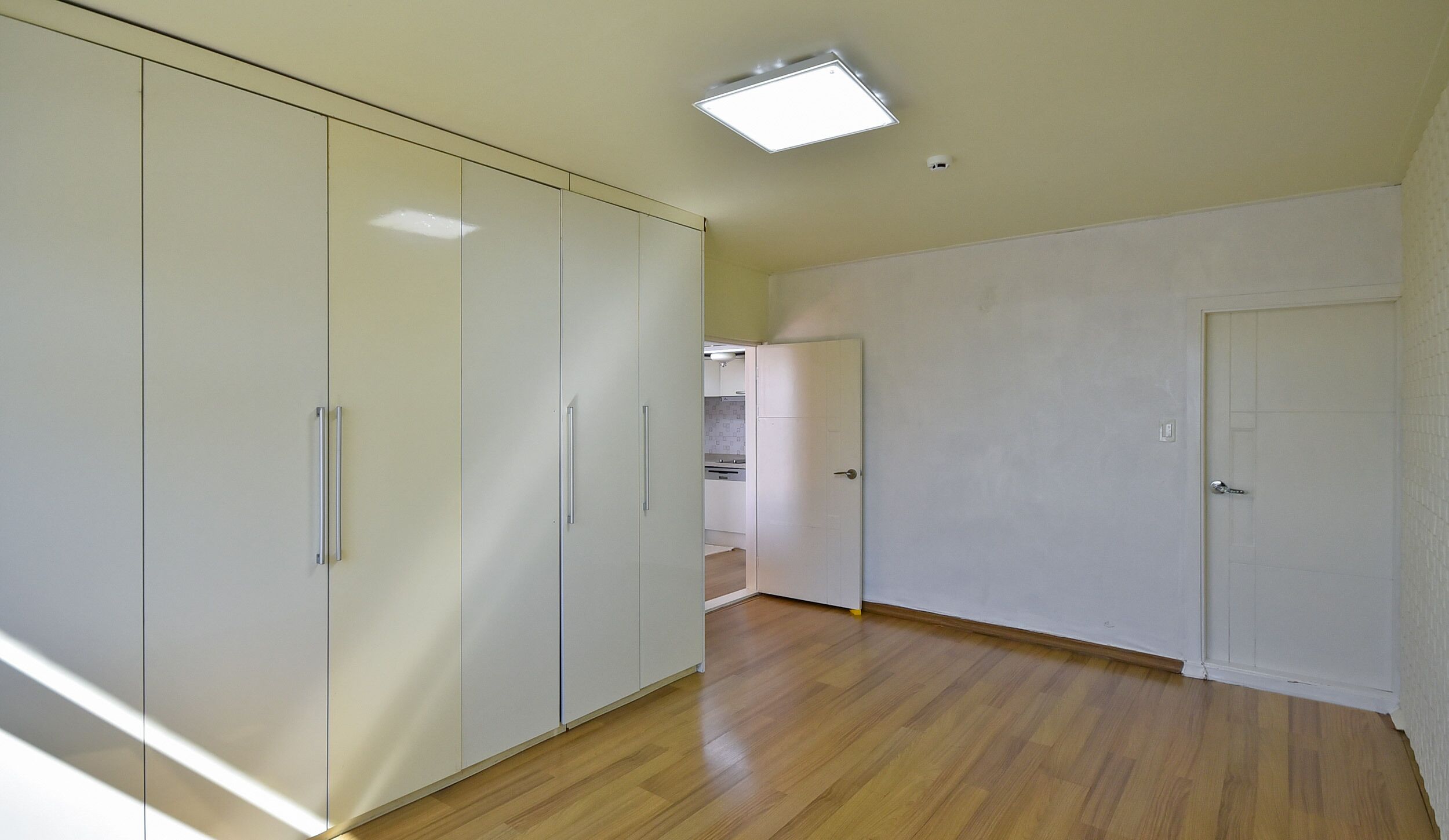 A room with a wooden floor, white walls, and a ceiling light. There is a door on the right and a wardrobe on the left.