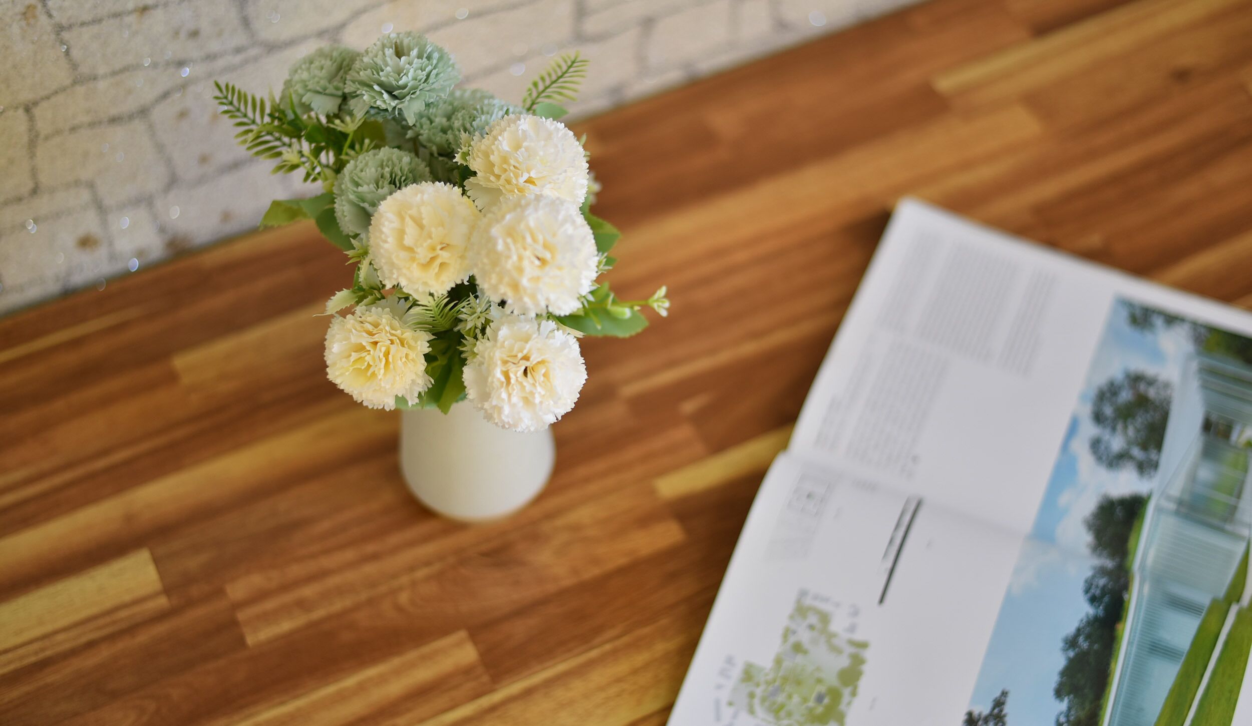 Un vase blanc orné de fleurs blanches et de feuilles vertes, posé sur une surface en bois, à côté d’un magazine.