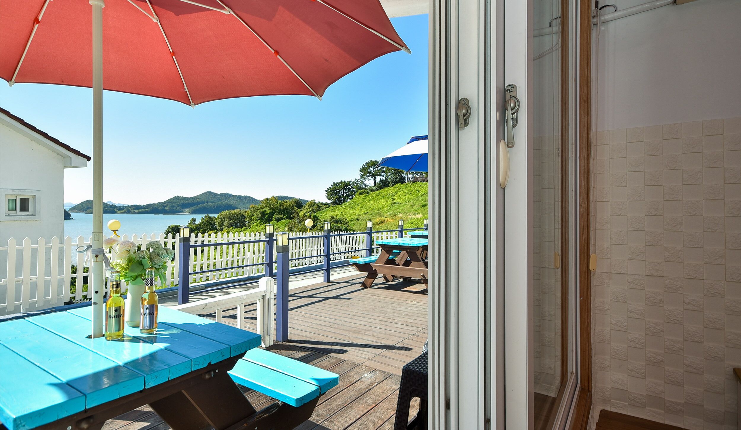 A wooden deck with a red umbrella, a table with bottles, and a view of the sea and hills.