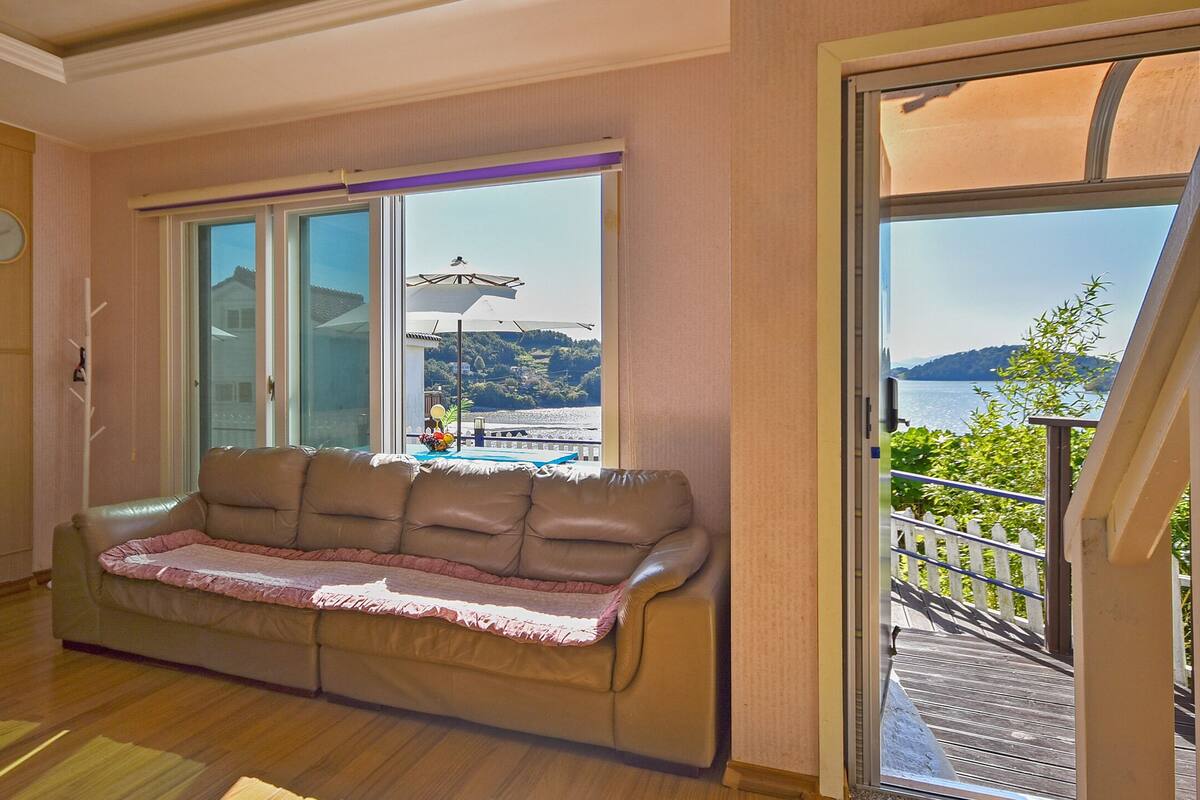 Ein gemütliches Wohnzimmer mit Sofa, Holzboden, großem Fenster mit Seeblick und Hügeln sowie einem Balkon mit Geländer.