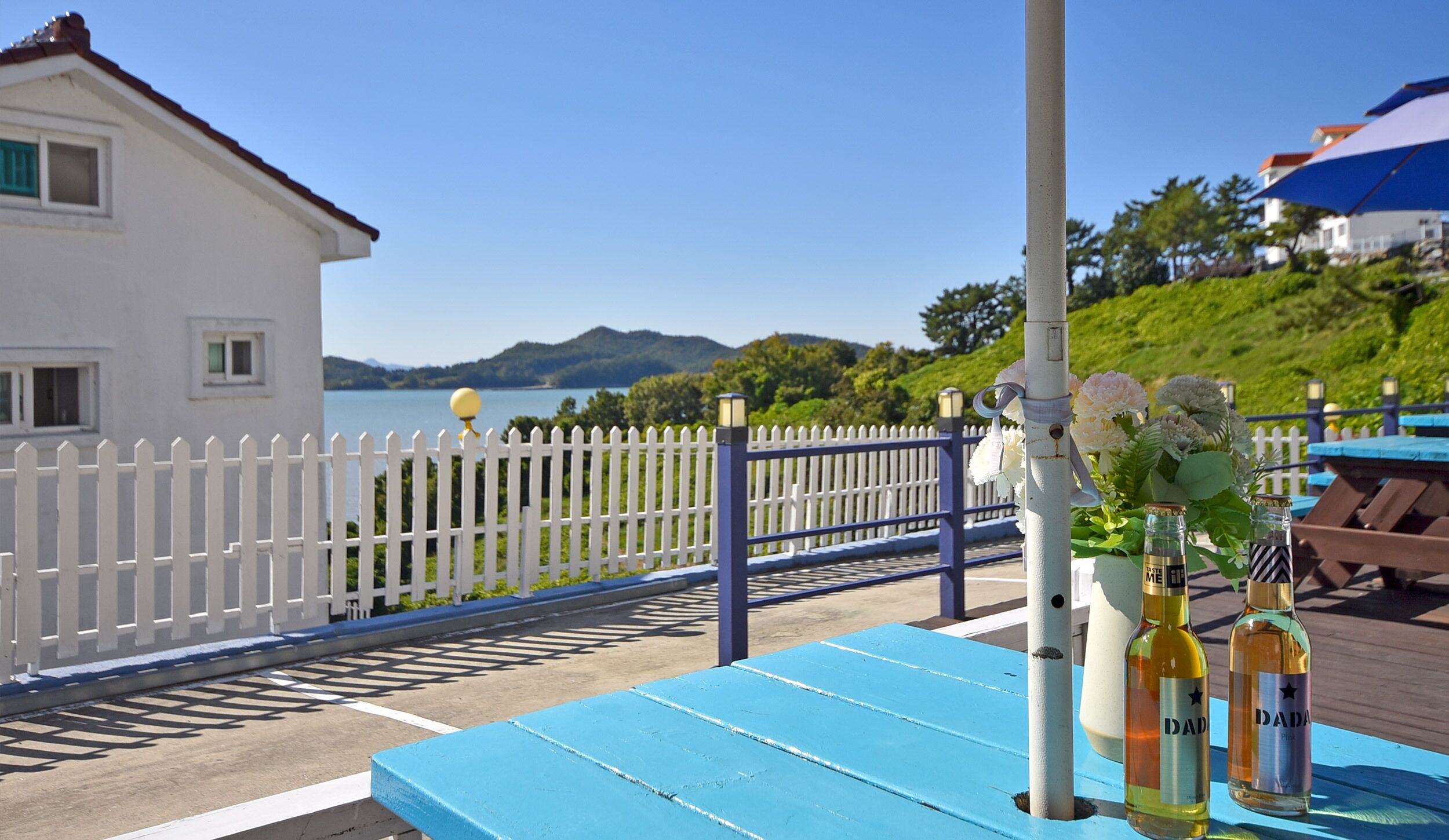 A coastal view with a white building, a white picket fence, and a blue table with bottles.