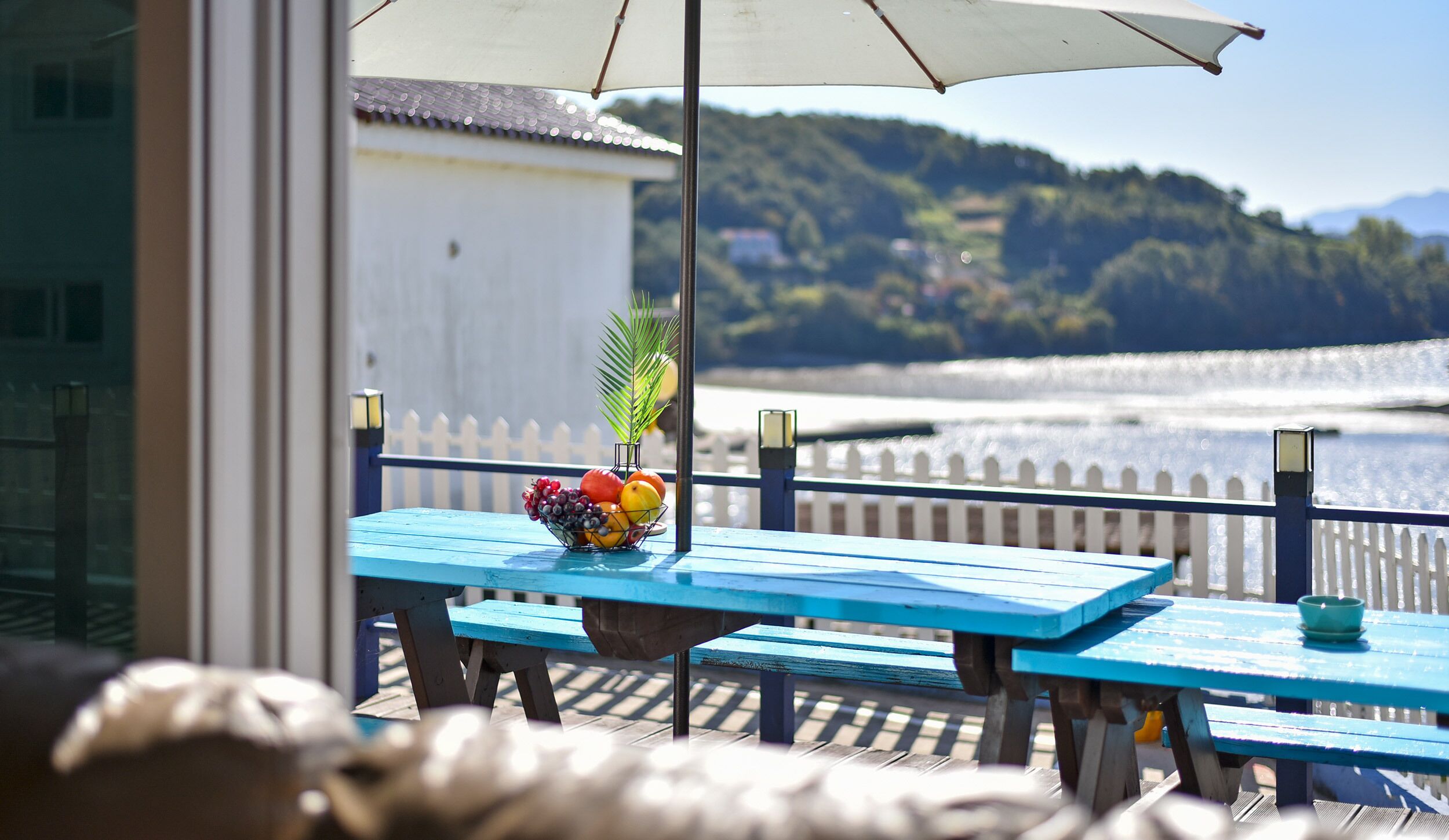 A blue table with a fruit bowl and a potted plant, overlooking a waterfront with a white fence and hills in the background.