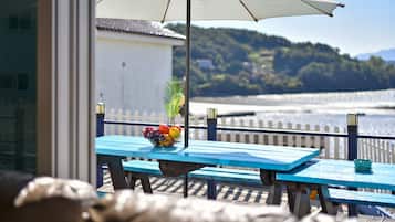 A blue table with a fruit bowl and a potted plant, overlooking a waterfront with a white fence and hills in the background.