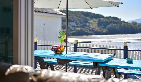 A blue table with a fruit bowl and a potted plant, overlooking a waterfront with a white fence and hills in the background.
