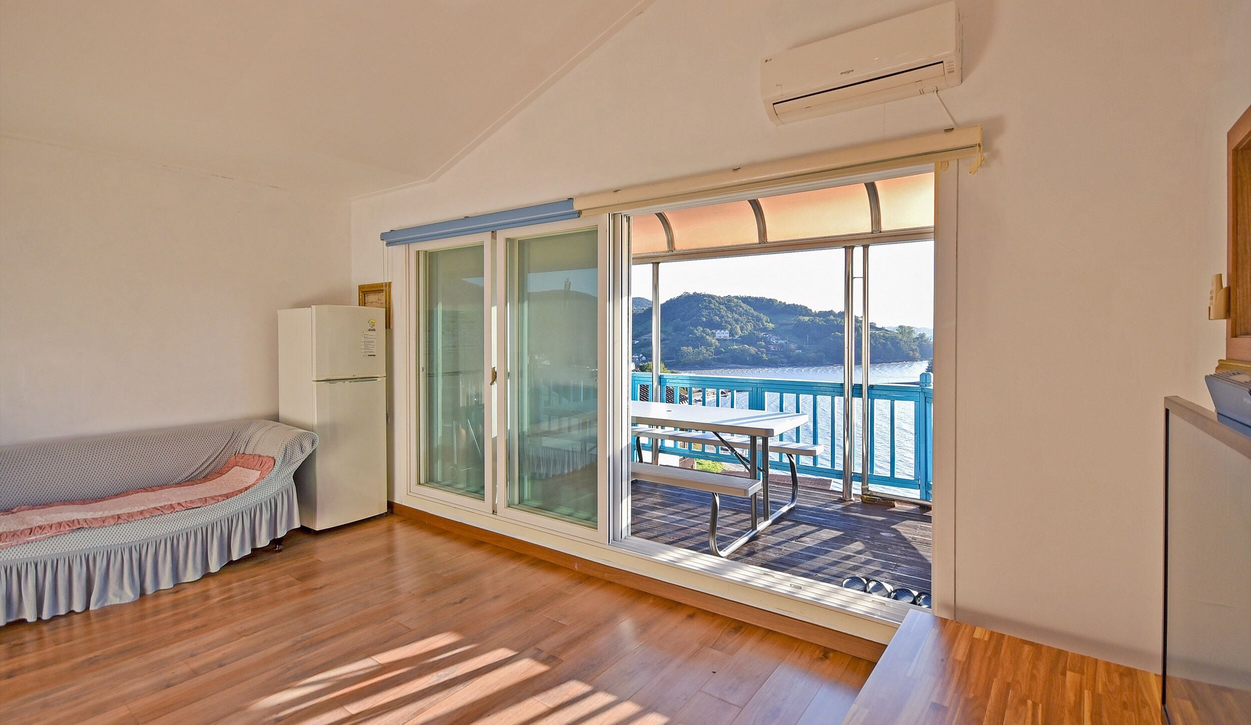 A room with a bed, a refrigerator, a sliding glass door leading to a balcony with a table and chairs, and a view of a body of water and hills.