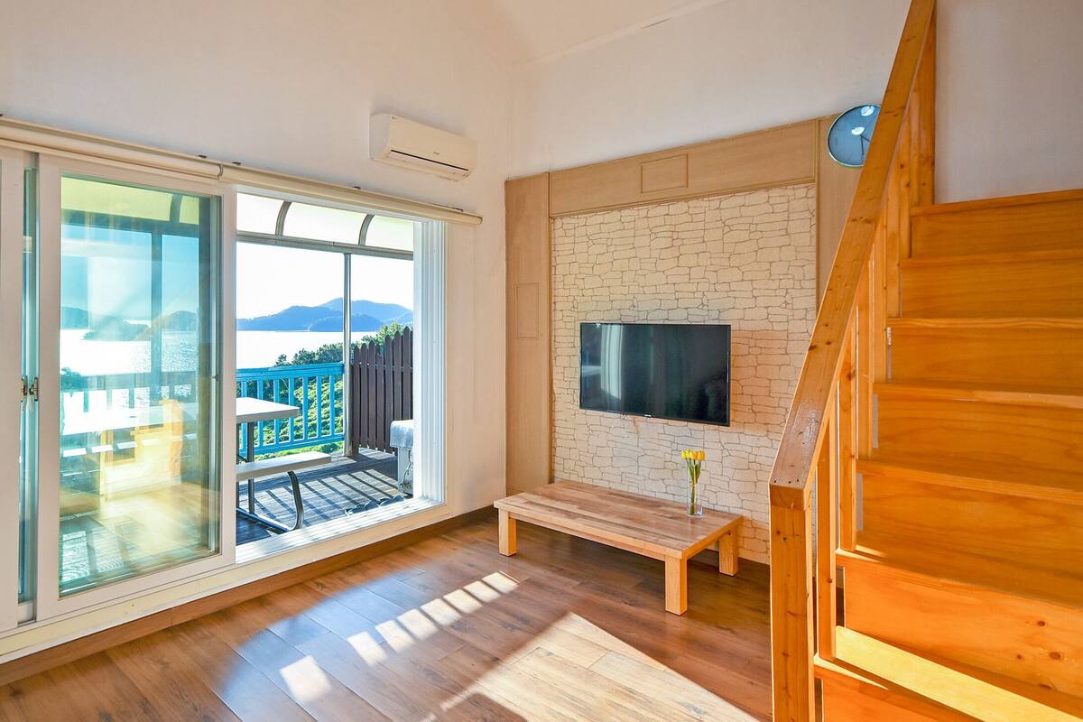 Ein modernes Zimmer mit einer großen Schiebeglasdoor, die auf einen Balkon mit Blick auf das Meer und die Berge führt. Dort steht eine Holzbank und ein Flachbildfernseher ist an einer Ziegelwand angebracht.