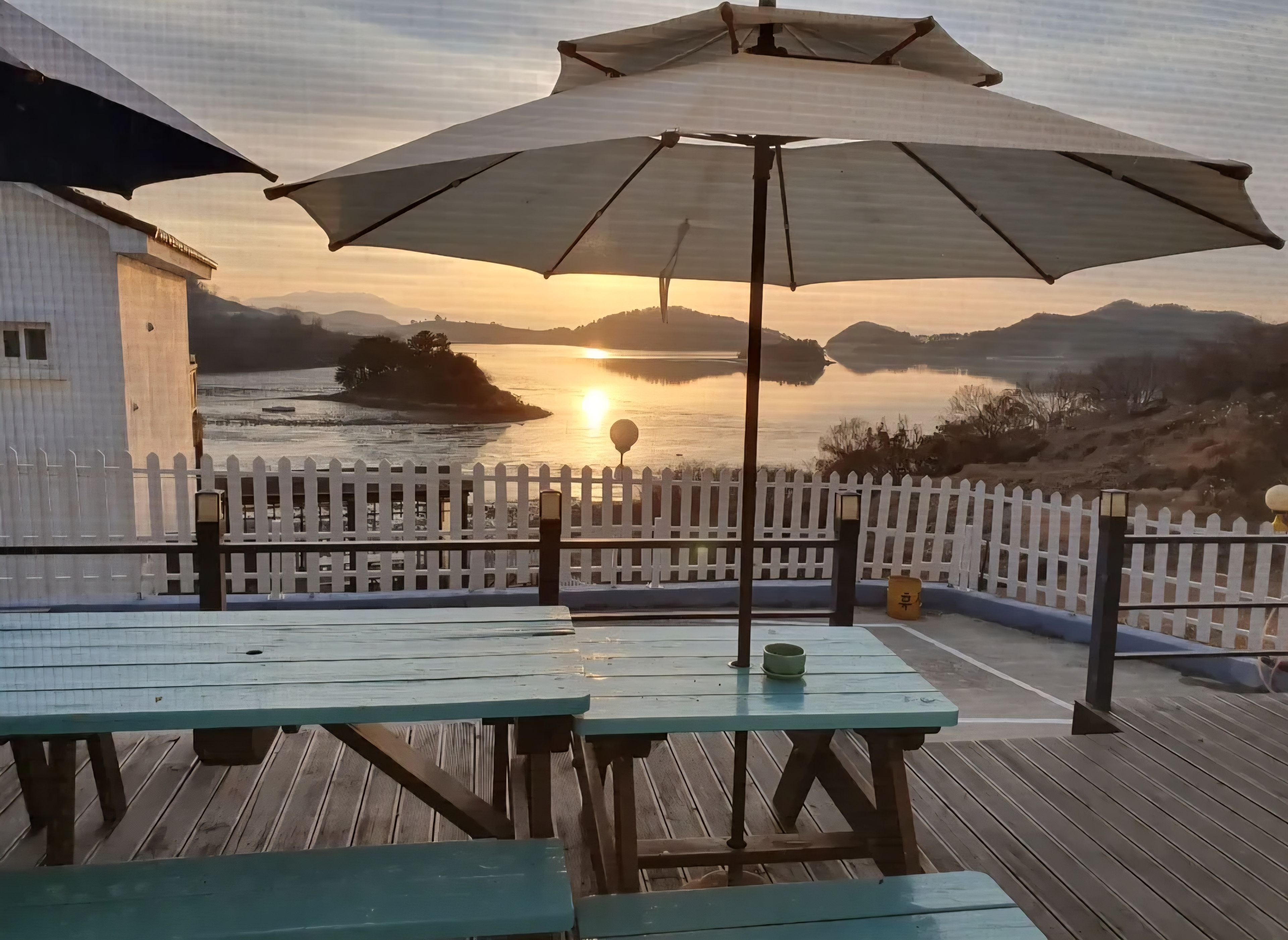 Une terrasse en bois avec des tables de pique-nique et un parasol, donnant sur une étendue d’eau au coucher du soleil.