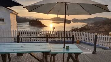 Une terrasse en bois avec des tables de pique-nique et un parasol, donnant sur une étendue d’eau au coucher du soleil.