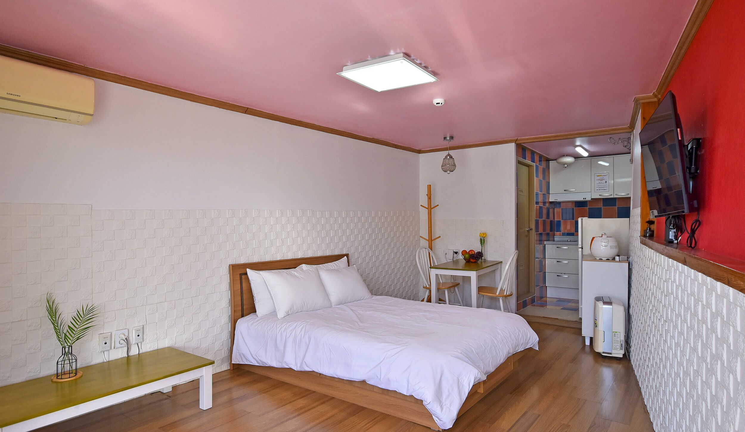 A bedroom with a bed, bedside table, and a small table with a chair. There is a kitchenette with a refrigerator, stove, and microwave.