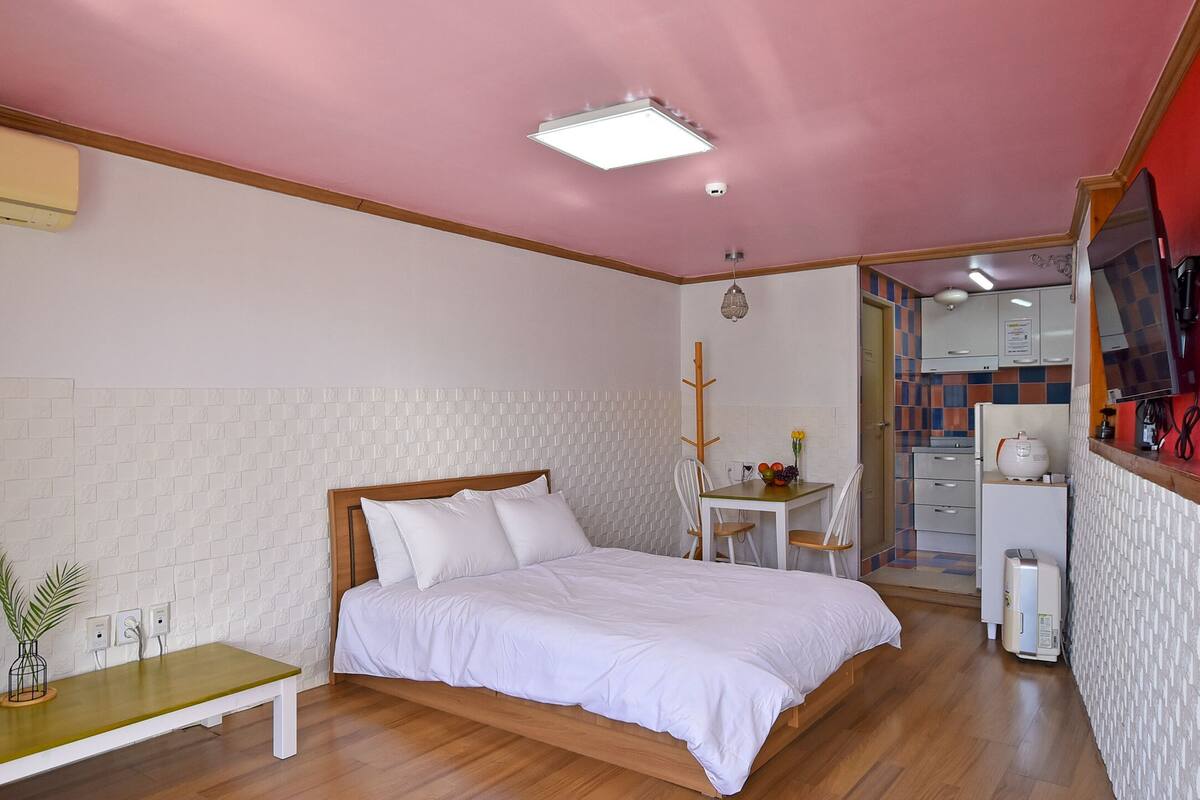 Ein Schlafzimmer mit Bett, Nachttisch und einem kleinen Tisch mit Stuhl. Es gibt eine Küchenzeile mit Kühlschrank, Herd und Mikrowelle.