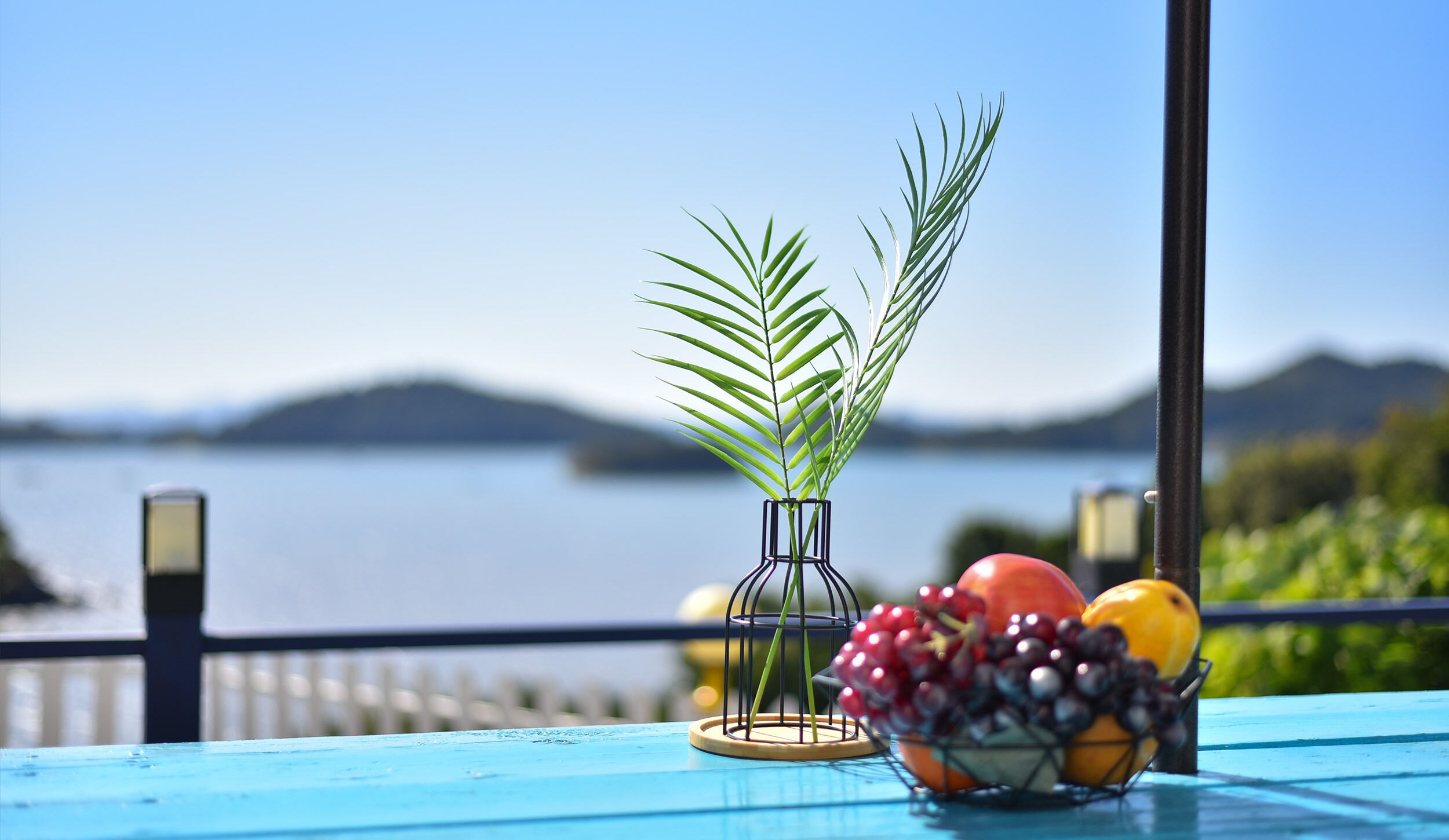 A table with a clear view of a lake and mountains in the background, a vase with a plant, and a bowl of fruit.