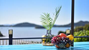 A table with a clear view of a lake and mountains in the background, a vase with a plant, and a bowl of fruit.