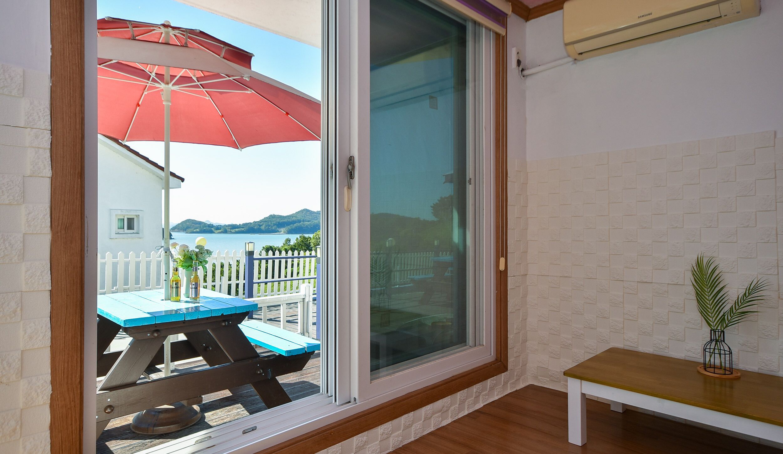 A room with a sliding glass door leading to a balcony with a red umbrella, a wooden table, and a bench.