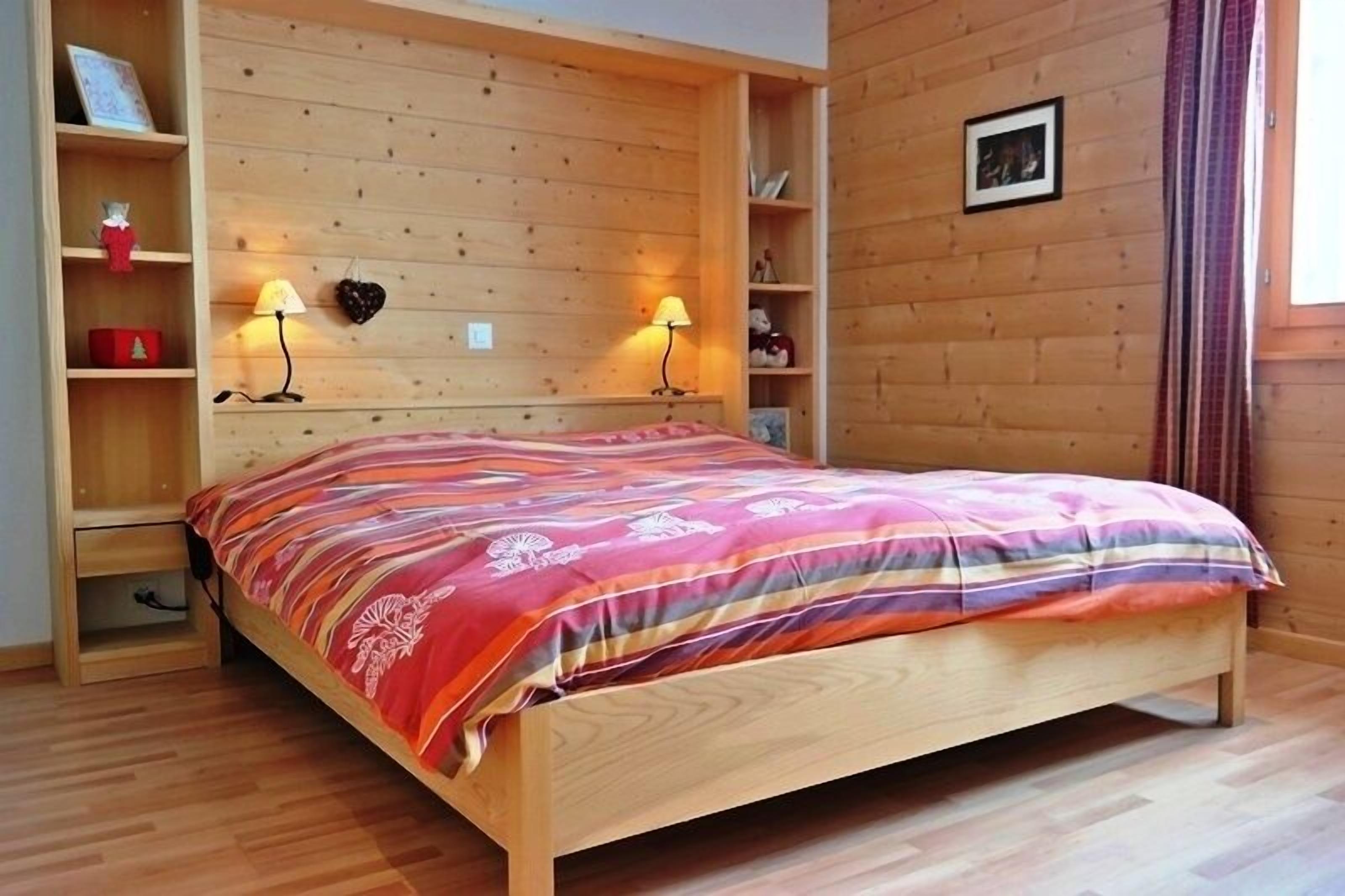 A wooden-paneled room with a bed, two wall-mounted lamps, and a shelf with decorative items.