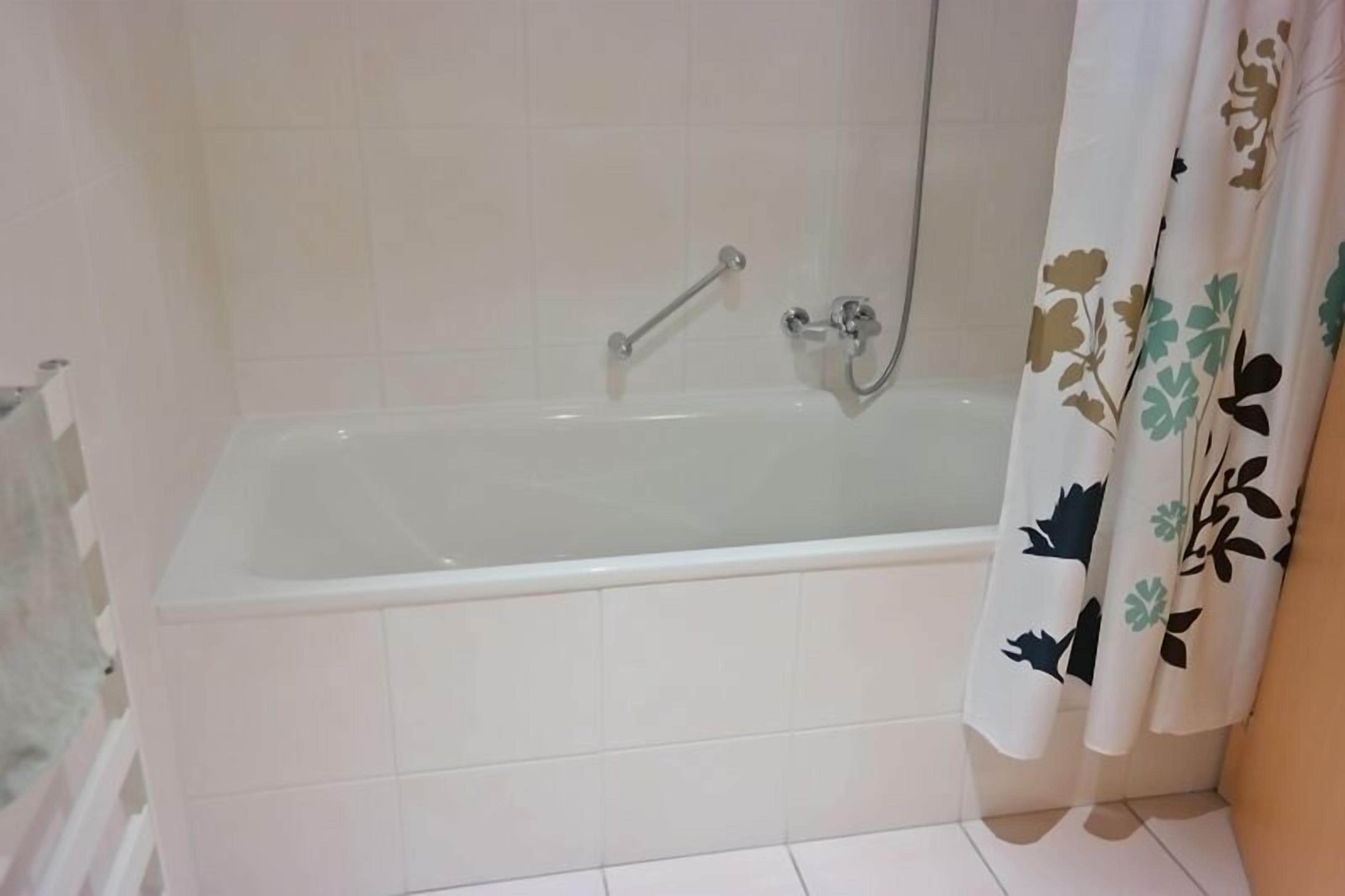 A standard bathtub with a handheld showerhead and a grab bar, set against a plain tiled wall.