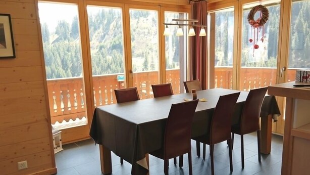 A dining area with a table set for six, large windows with a view of a forested mountain, and a wooden balcony railing. - Appartement Moilles 15 (Val d'Illiez)