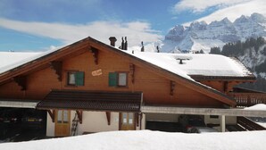 A wooden chalet with a sloped roof, a wooden porch, and a view of snow-covered mountains in the background. - Appartement Moilles 15 (Val d'Illiez)