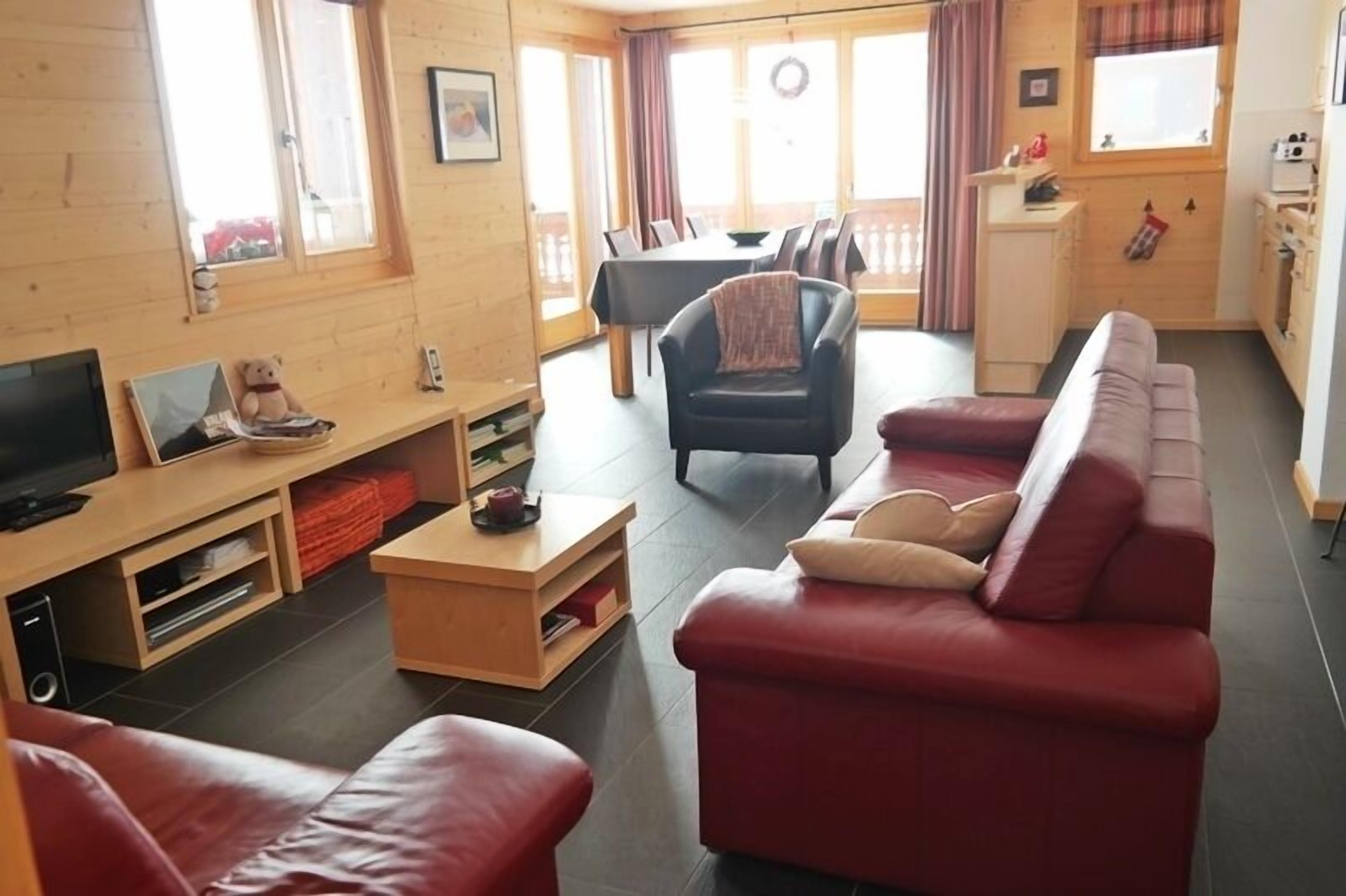 A cozy living room with a red sofa, a wooden coffee table, and a TV. There is a dining area with a table and chairs, and a kitchen area with a stove and cabinets.