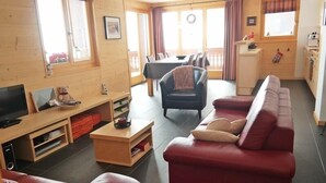 A cozy living room with a red sofa, a wooden coffee table, and a TV. There is a dining area with a table and chairs, and a kitchen area with a stove and cabinets. - Appartement Moilles 15 (Val d'Illiez)