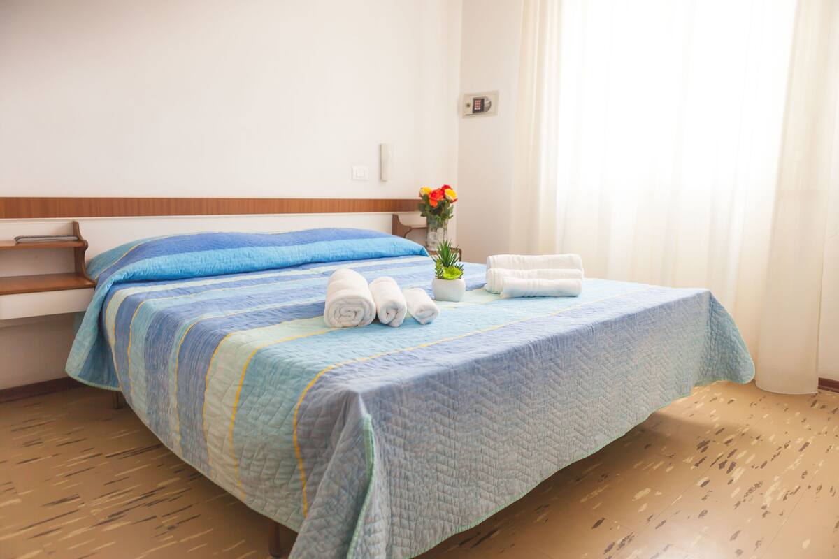 Een net opgemaakt bed met een blauw-witte streepdessin, een vaas met bloemen, en opgerolde handdoeken op het bed.