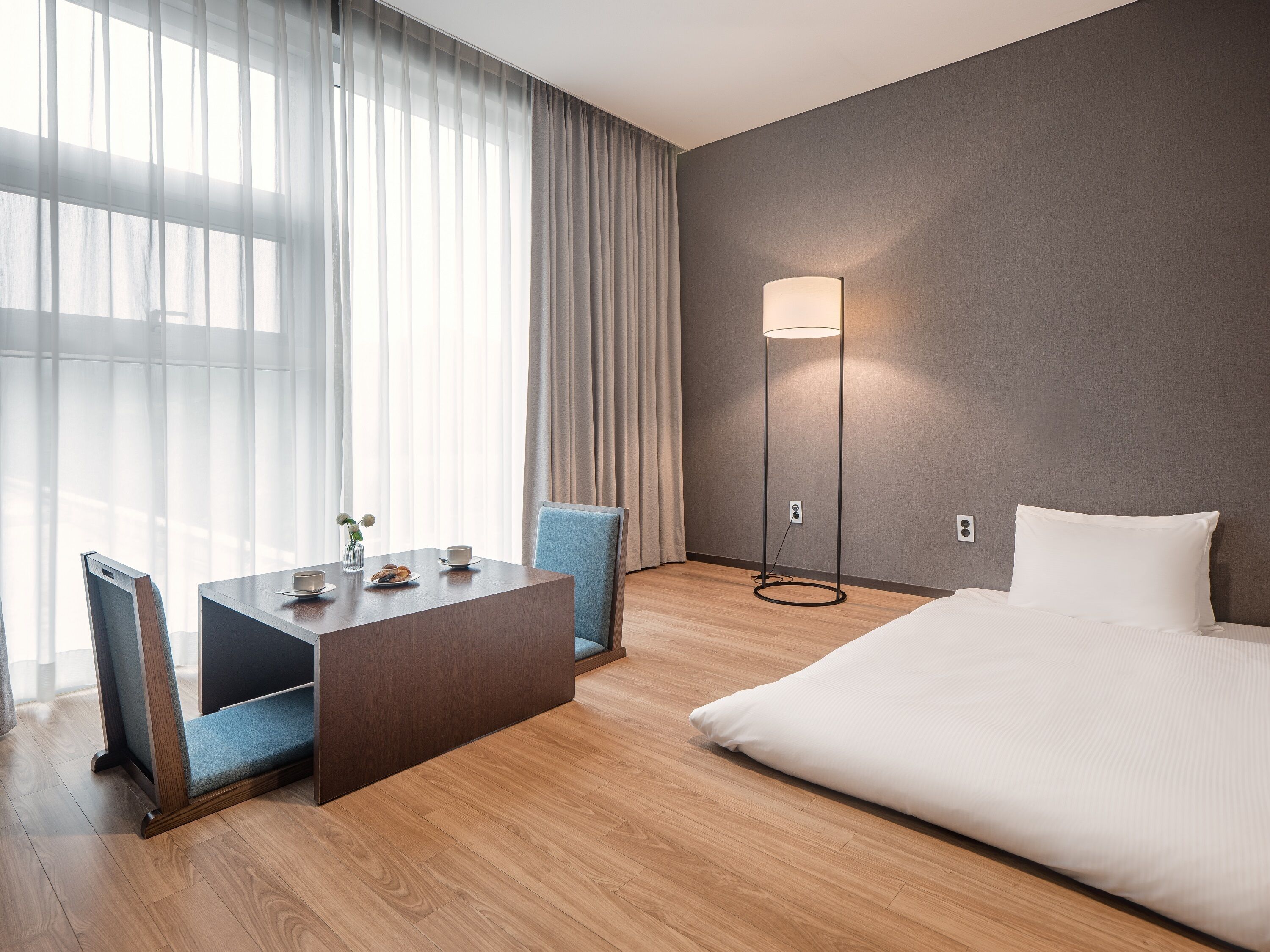 Une chambre d’hôtel avec un lit, un bureau avec des chaises, une lampe sur pied et une grande fenêtre avec des rideaux.