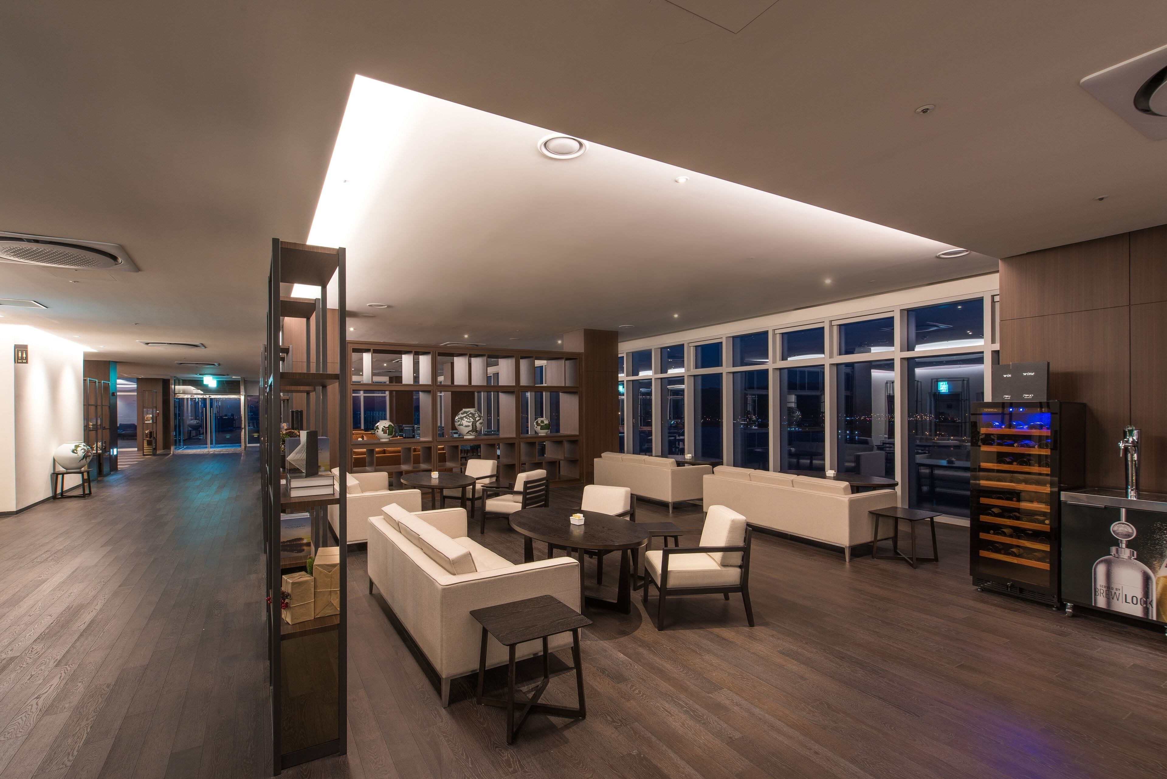 A modern hotel lobby with a wooden floor, white sofas, and a bar area with a wine cooler.