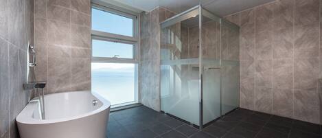 A modern bathroom with a freestanding bathtub, a glass shower enclosure, and a large window offering a view of the sea.