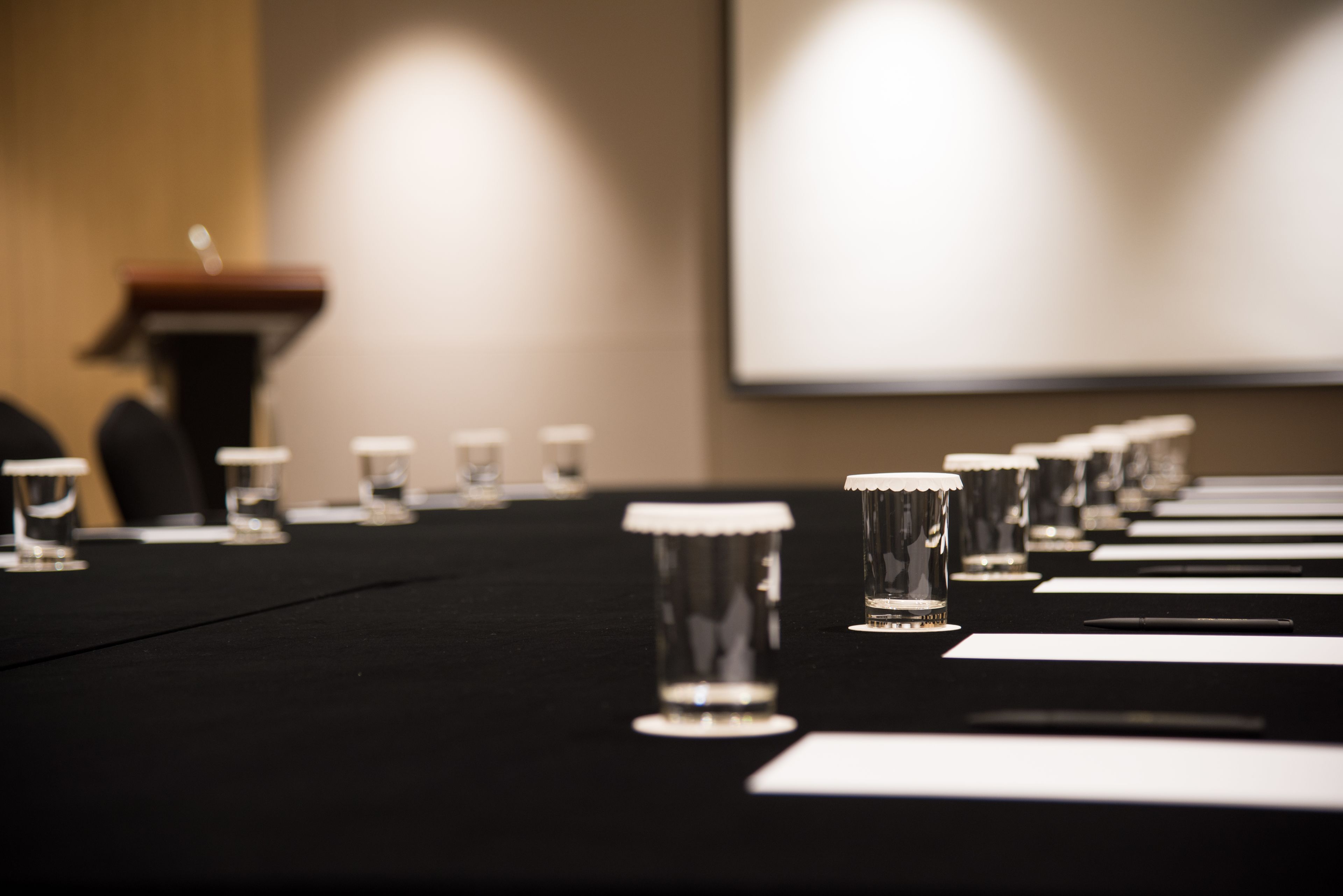 Una mesa de conferencias con vasos y papeles, un atril y una pantalla de proyección al fondo.