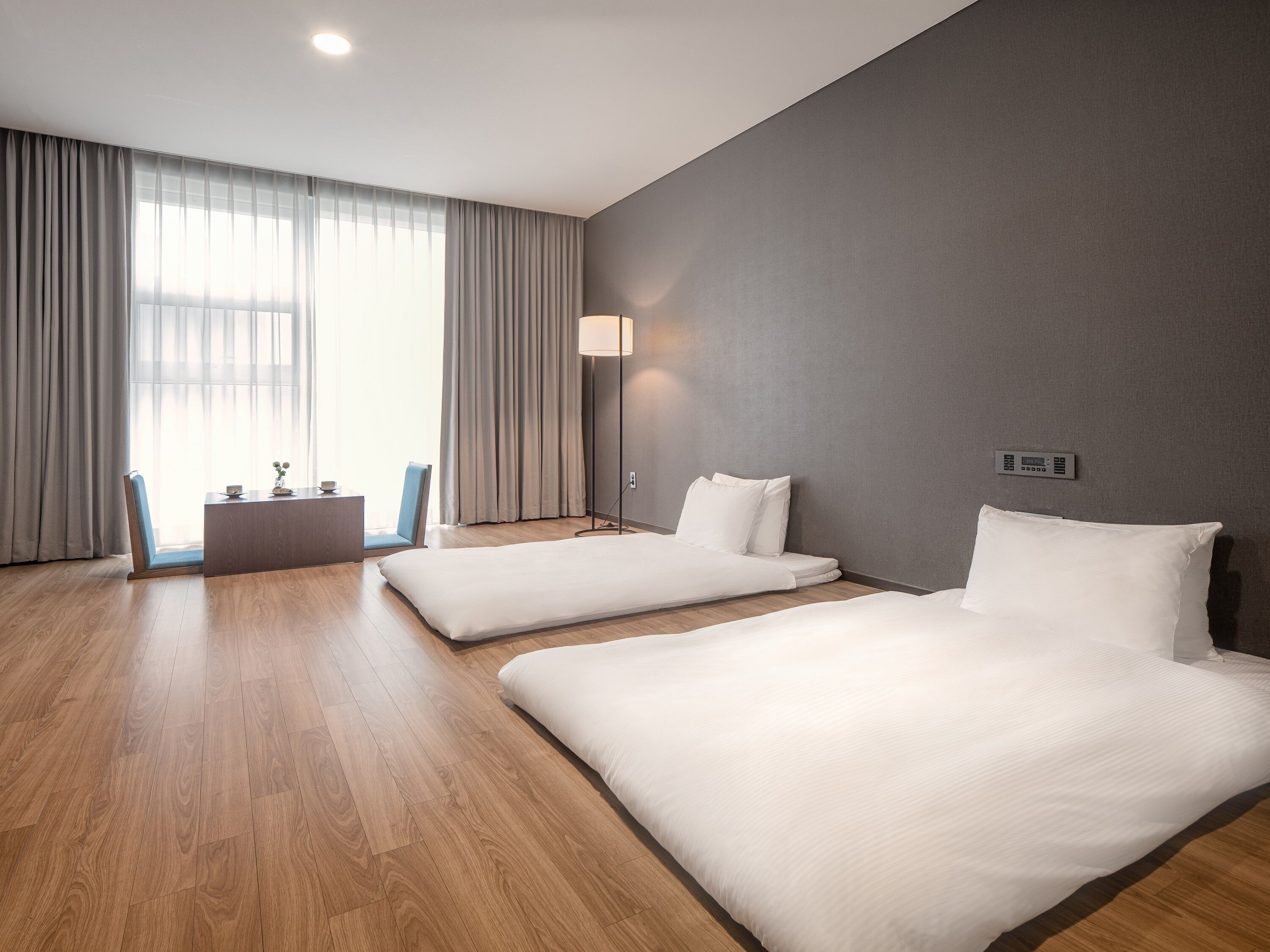 A modern hotel room with two single beds, a desk, a chair, and a large window with curtains.