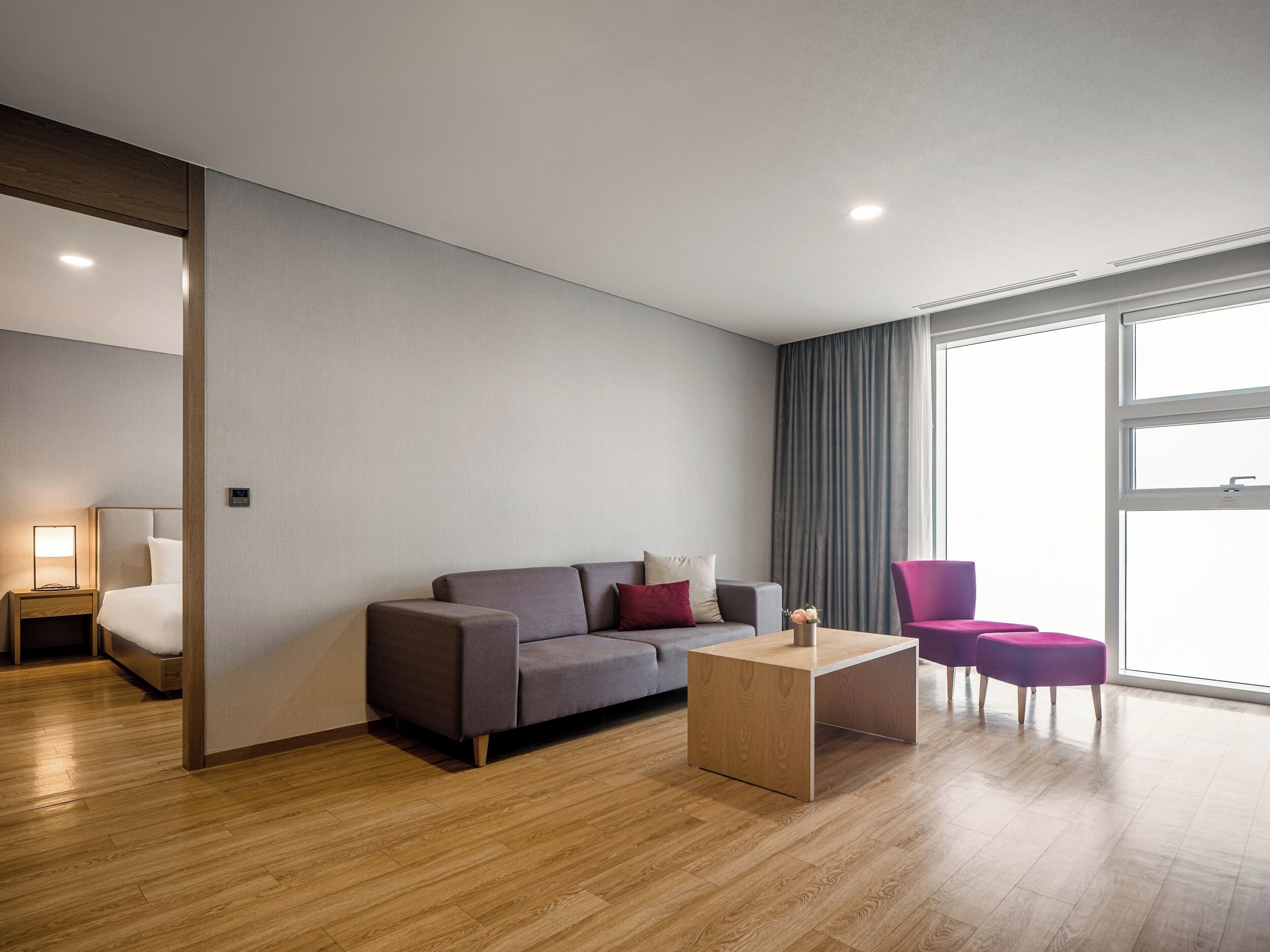 Una habitación de hotel moderna con un sofá gris, una mesa de centro de madera y un sillón morado.
