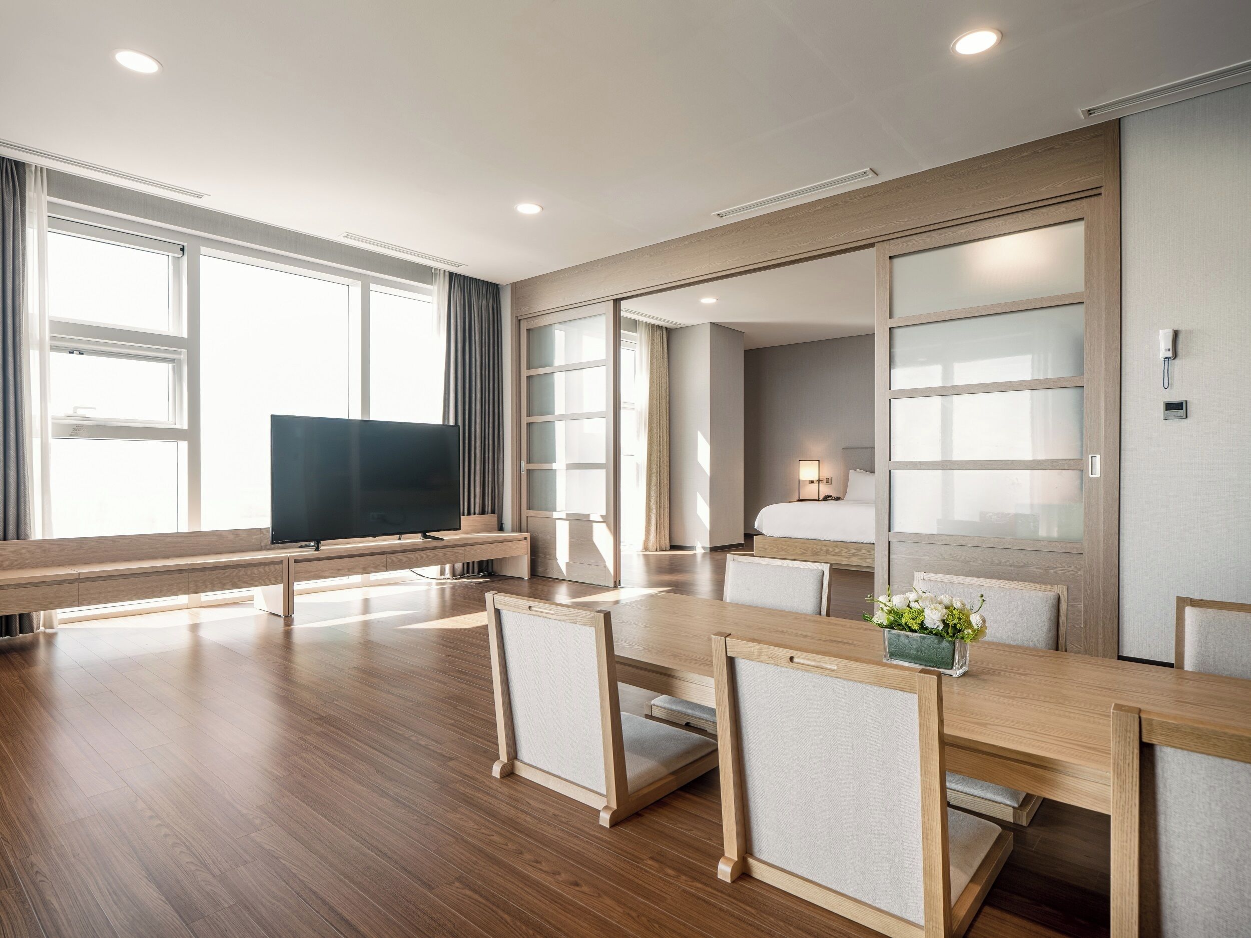 A modern hotel room with a large flat-screen TV, a bed with a bedside table, a desk with a chair, and large windows.