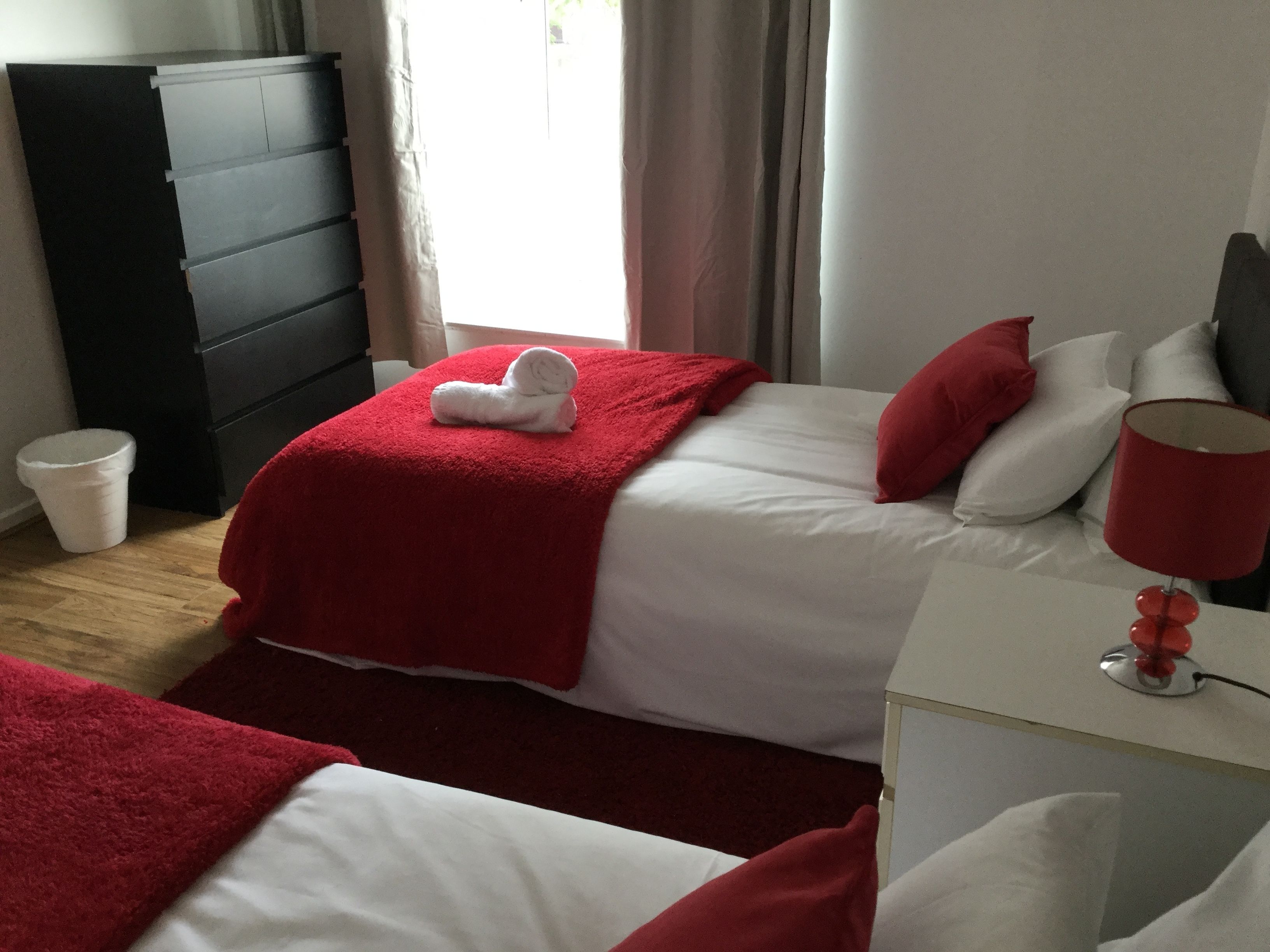 Et hotelværelse med to senge, begge med hvide sengetæpper og røde detaljer, en rød lampe på et natbord og en sort garderobe.