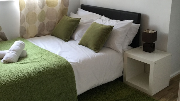 A neatly made bed with white linens and green pillows, a bedside table with a lamp, and a framed picture on the wall. - Nice 4 Bedroom near Basildon Town Center (Basildon)