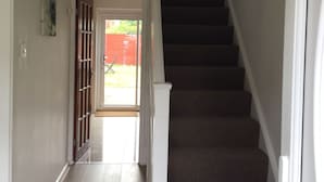 A hallway with a wooden floor, a staircase with dark carpeted steps, and a green wall lamp. - Nice 4 Bedroom near Basildon Town Center (Basildon)