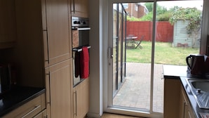 A kitchen with wooden cabinets, a black countertop, and a sliding glass door leading to a patio with a table and chairs. - Nice 4 Bedroom near Basildon Town Center (Basildon)