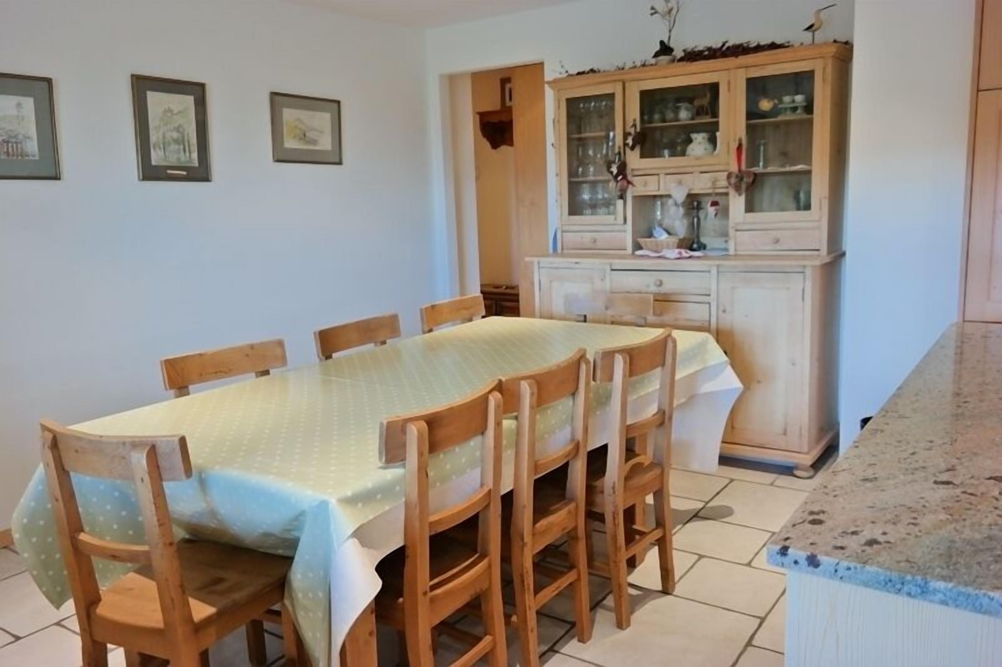 A dining area with a table covered by a green tablecloth, surrounded by wooden chairs. There is a wooden cabinet with glass doors in the background.