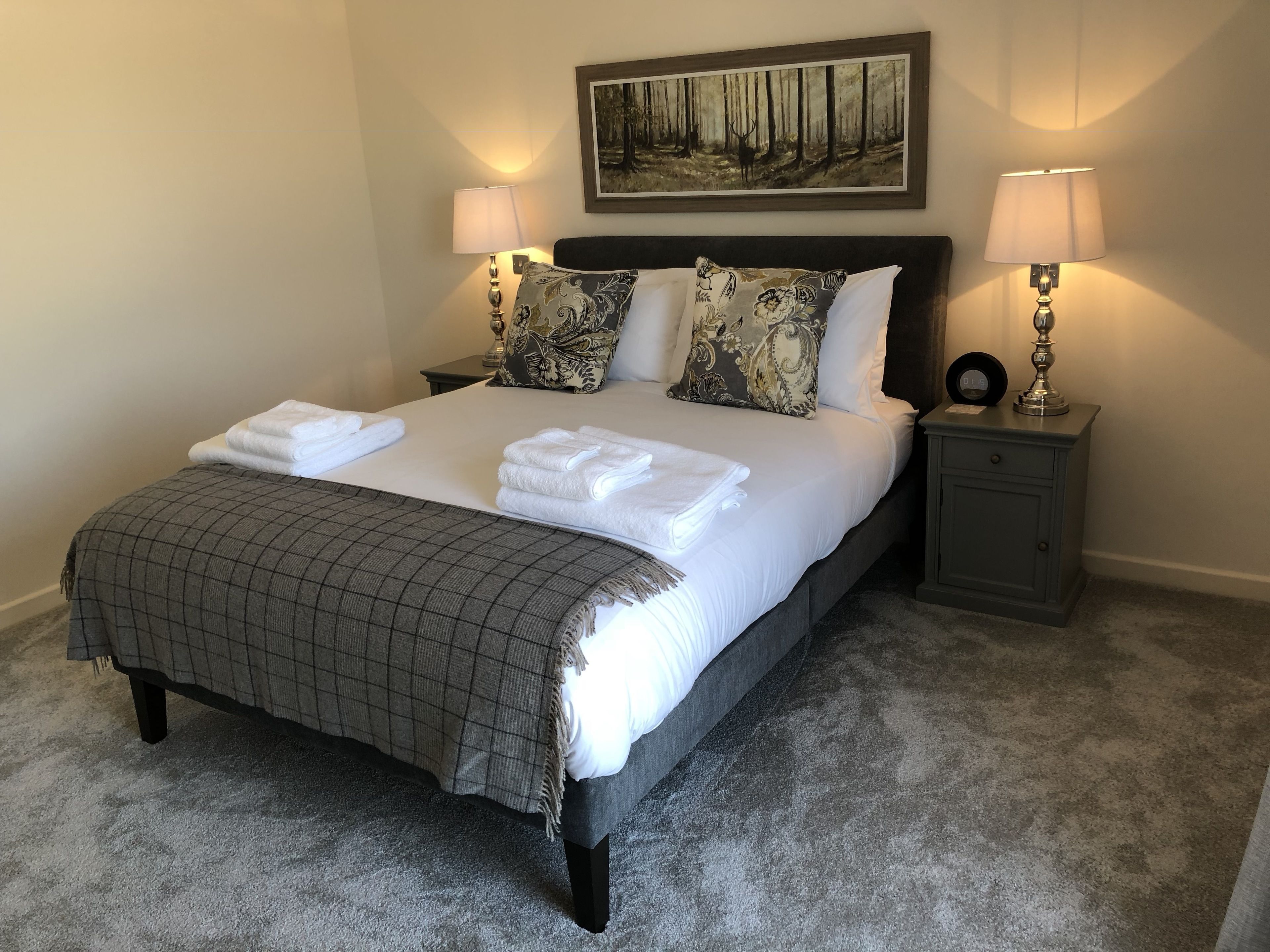 A neatly made bed with white linens and a grey checkered throw, flanked by two bedside tables with lamps, a framed forest painting above the bed, and a grey carpeted floor.