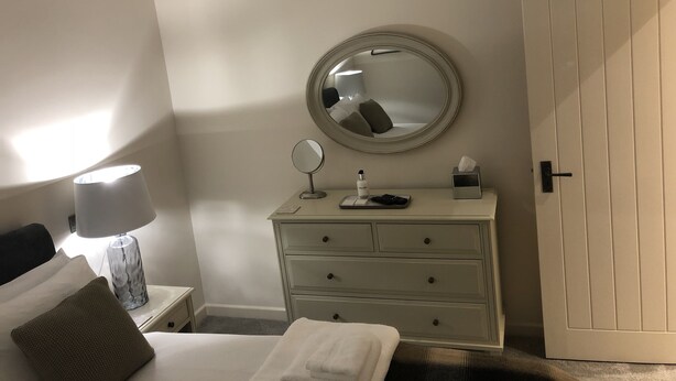 Una camera d'albergo con un letto, un comodino con una lampada, una cassettiera e uno specchio, oltre a una porta.