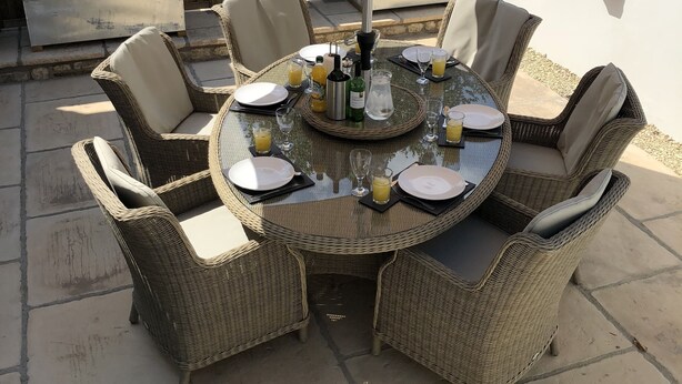 A round outdoor dining table with wicker chairs, an umbrella, and a planter with flowers in the background. - Camerton Inn Cottage (Bath)