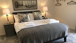 A neatly made bed with a checkered blanket, two pillows with floral patterns, and two bedside lamps. - Camerton Inn Cottage (Bath)