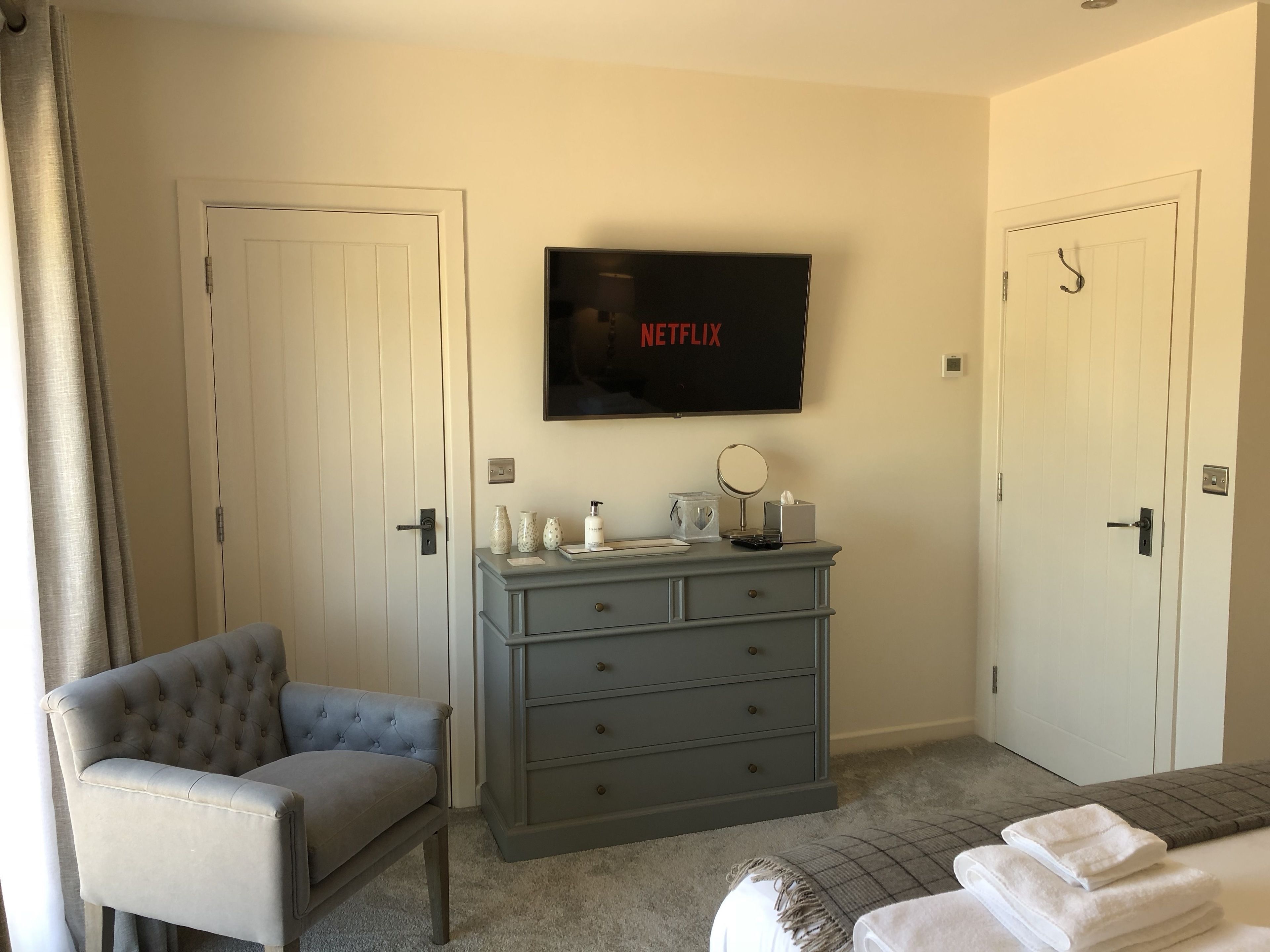 Una camera d'albergo con una poltrona imbottita grigia, un comò grigio con cassetti, un televisore a schermo piatto montato sopra il comò che mostra 'NETFLIX', e due porte chiuse.