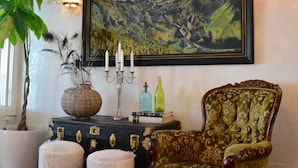 Una habitación con un cuadro de paisaje grande, un sillón tapizado en verde, un baúl antiguo y una planta en maceta.