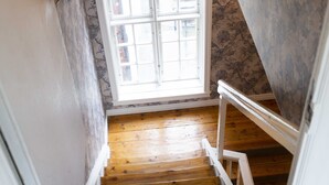 Una escalera de madera con barandales blancos que lleva a una habitación con papel tapiz estampado y un ventanal.