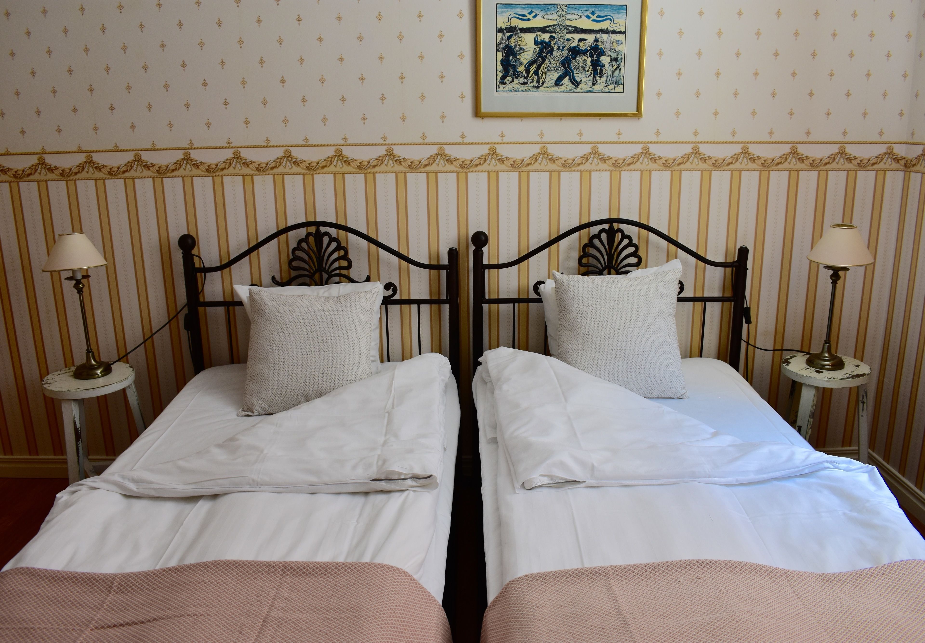 Una cama doble con sábanas blancas y dos almohadas, flanqueada por dos mesitas de noche con lámparas, en una habitación con papel tapiz a rayas y un cuadro enmarcado sobre la cama.